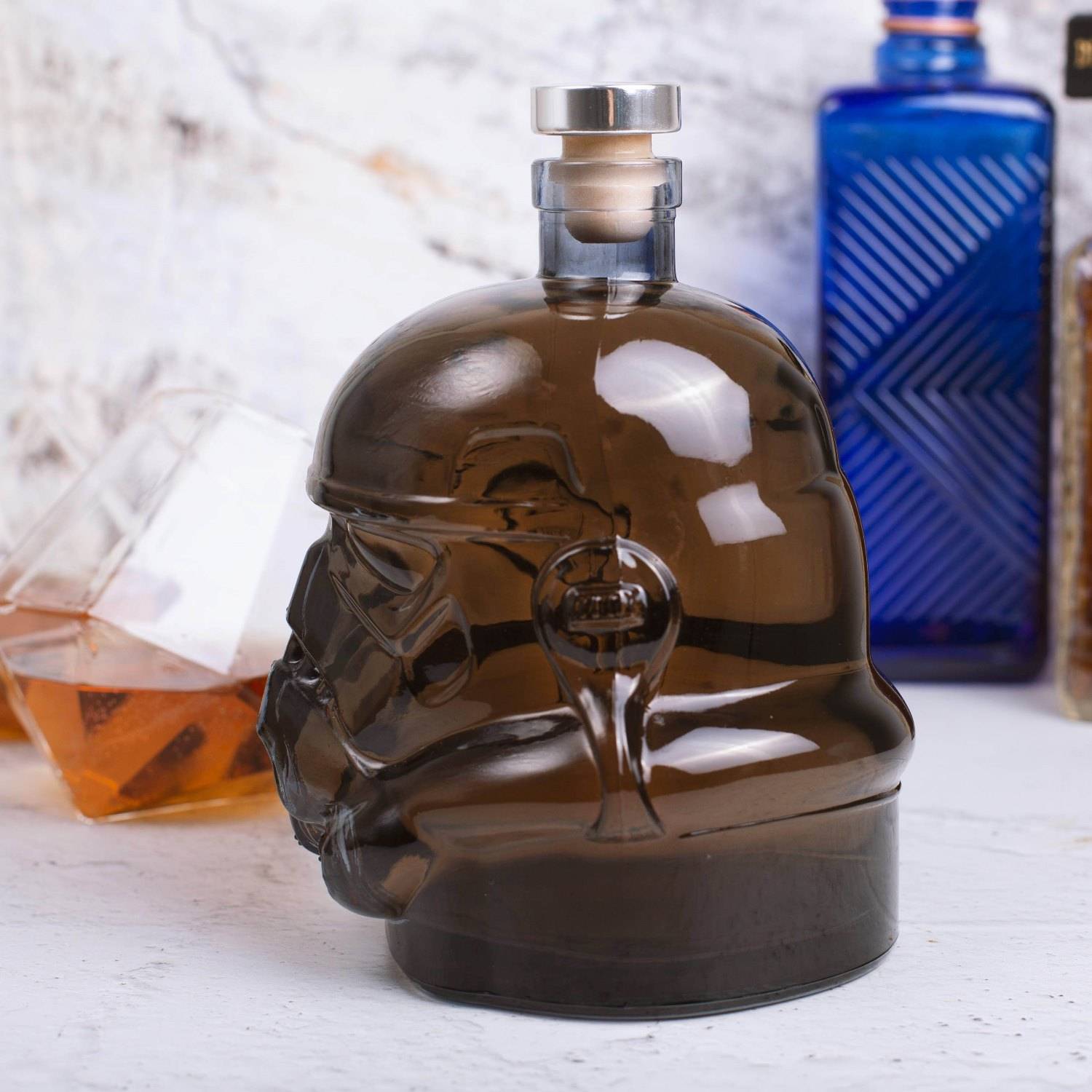 Dunkelschädel-förmige Likörflasche auf einer weißen Oberfläche, mit einem Glas Bernstein-Whiskey und einer blauen Flasche im Hintergrund.
