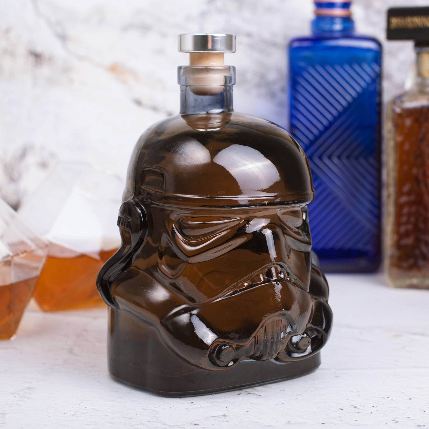 Stormtrooper-förmige Glasflasche gefüllt mit dunklem Schnaps auf einer weißen Oberfläche, mit anderen unscharfen Whiskey-Flaschen im Hintergrund.