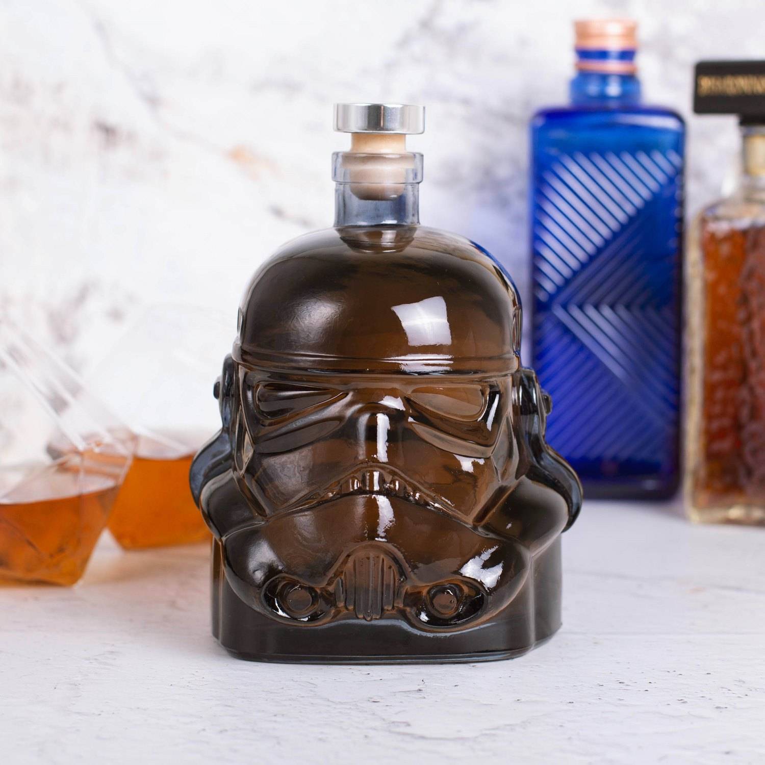Stormtrooper-förmige Glaslikörflasche gefüllt mit dunkelbrauner Flüssigkeit, zentriert auf einer weißen Oberfläche mit unscharfen Flaschen im Hintergrund.