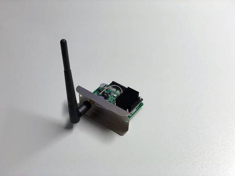 Kleine elektronische Leiterplatte in einem Metallgehäuse mit einer vertikalen Antenne, dargestellt auf weißem Hintergrund.