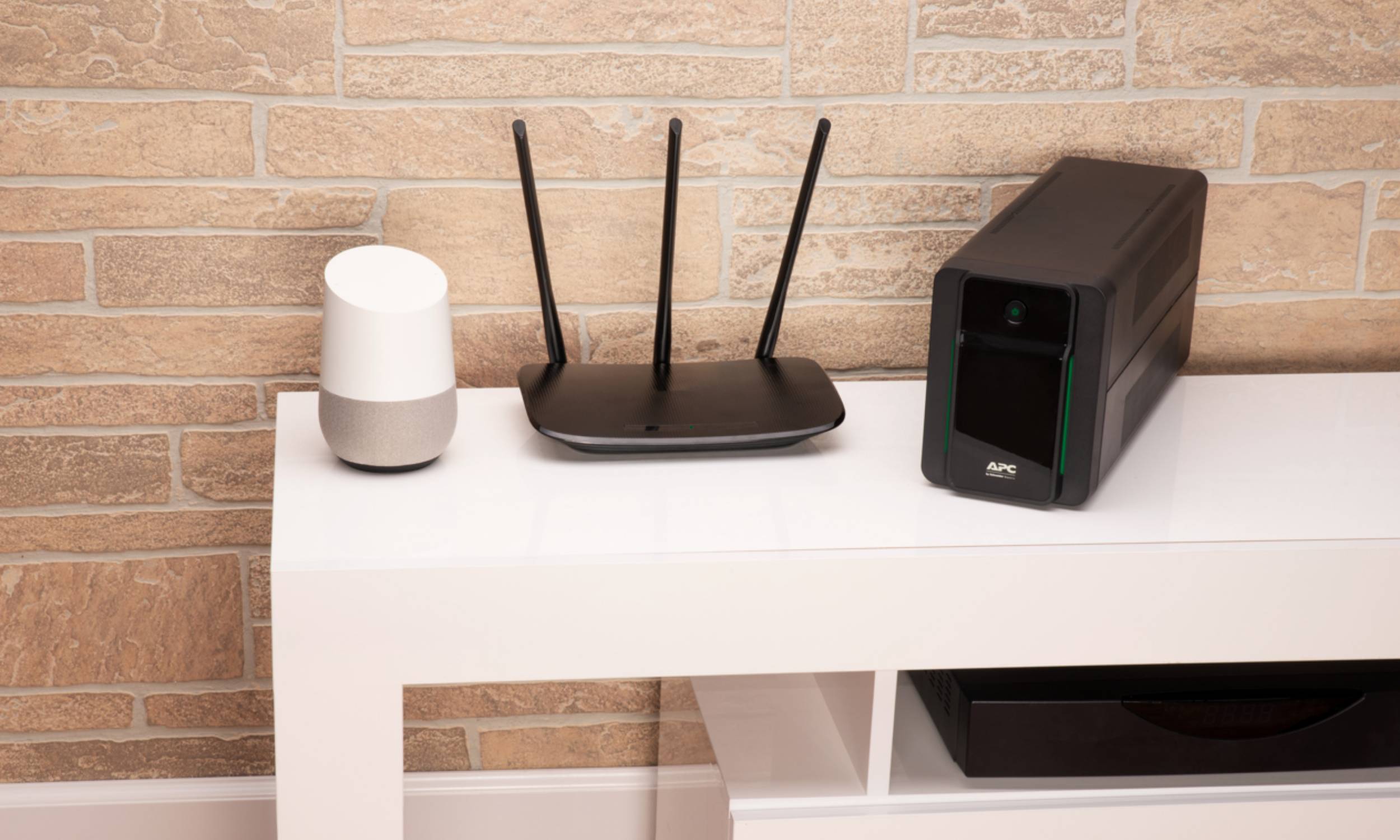 Weißer Tisch mit einem Smart Speaker, WLAN-Router und APC-Batterienotstromversorgung vor einer Backsteinmauer.