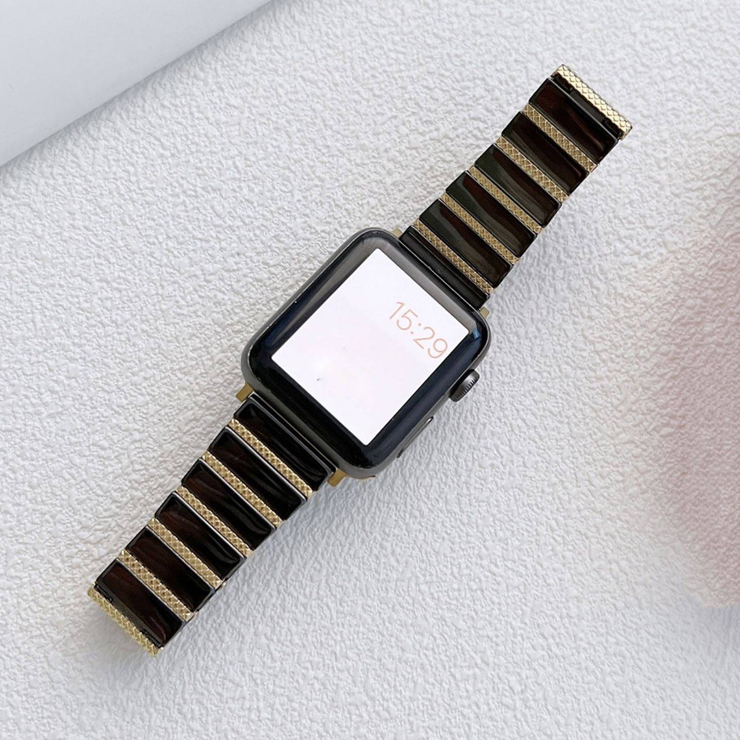 Eine schwarze rechteckige Smartwatch mit einem gestreiften braun-goldenen Armband auf hellem strukturiertem Hintergrund, Bildschirm zeigt 15:29 an.