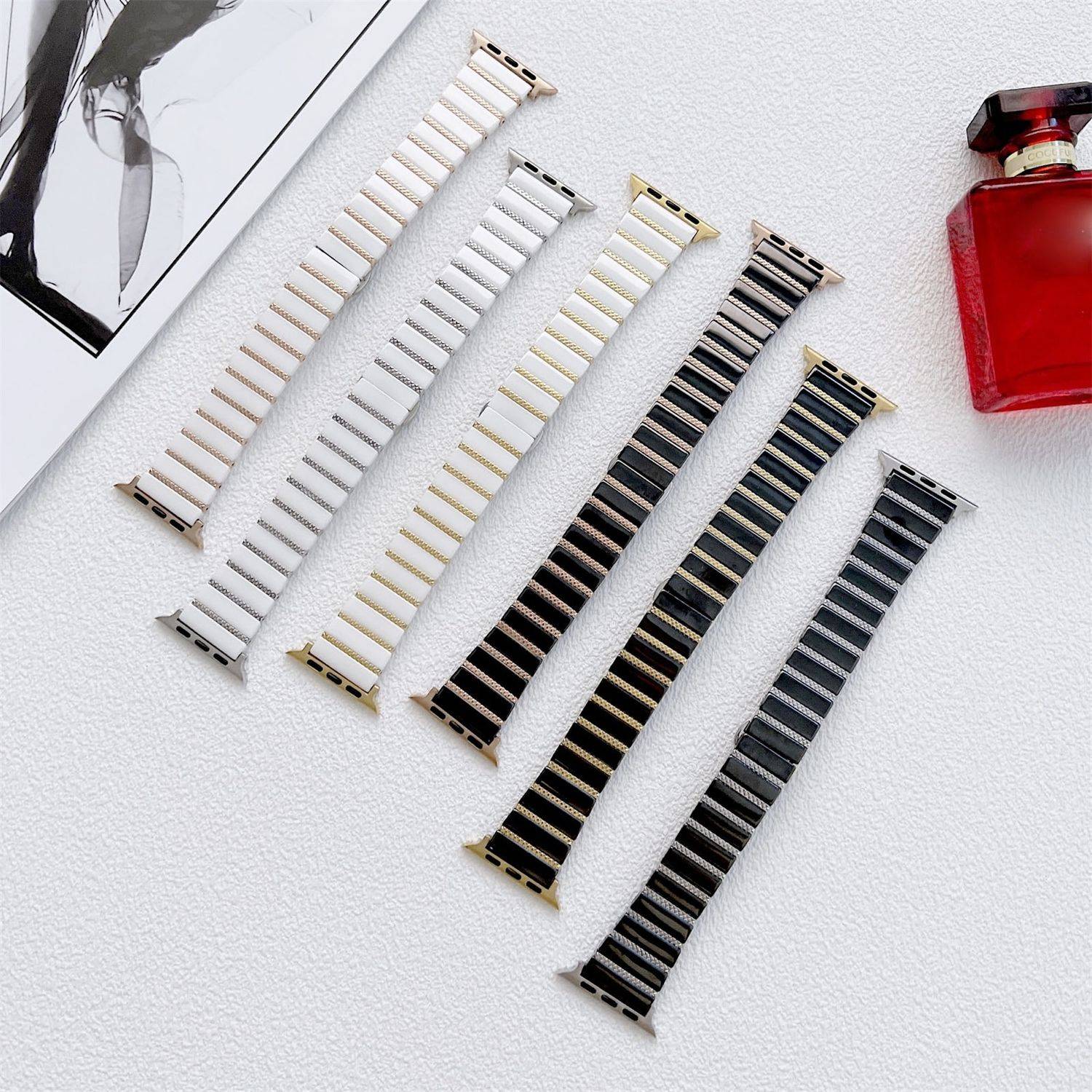 Sechs austauschbare Uhrenarmbänder diagonal auf einer weißen Oberfläche angeordnet, von weiß bis schwarz, neben einer roten Parfümflasche und einem Teil eines Modefotos.