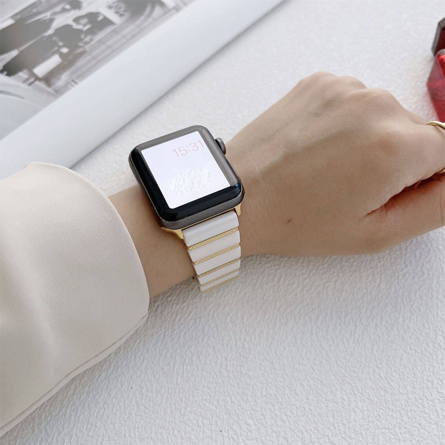 Nahaufnahme eines Handgelenks, das eine quadratische Smartwatch mit einem weiß-goldenen Armband auf einer leicht strukturierten hellen Oberfläche trägt.
