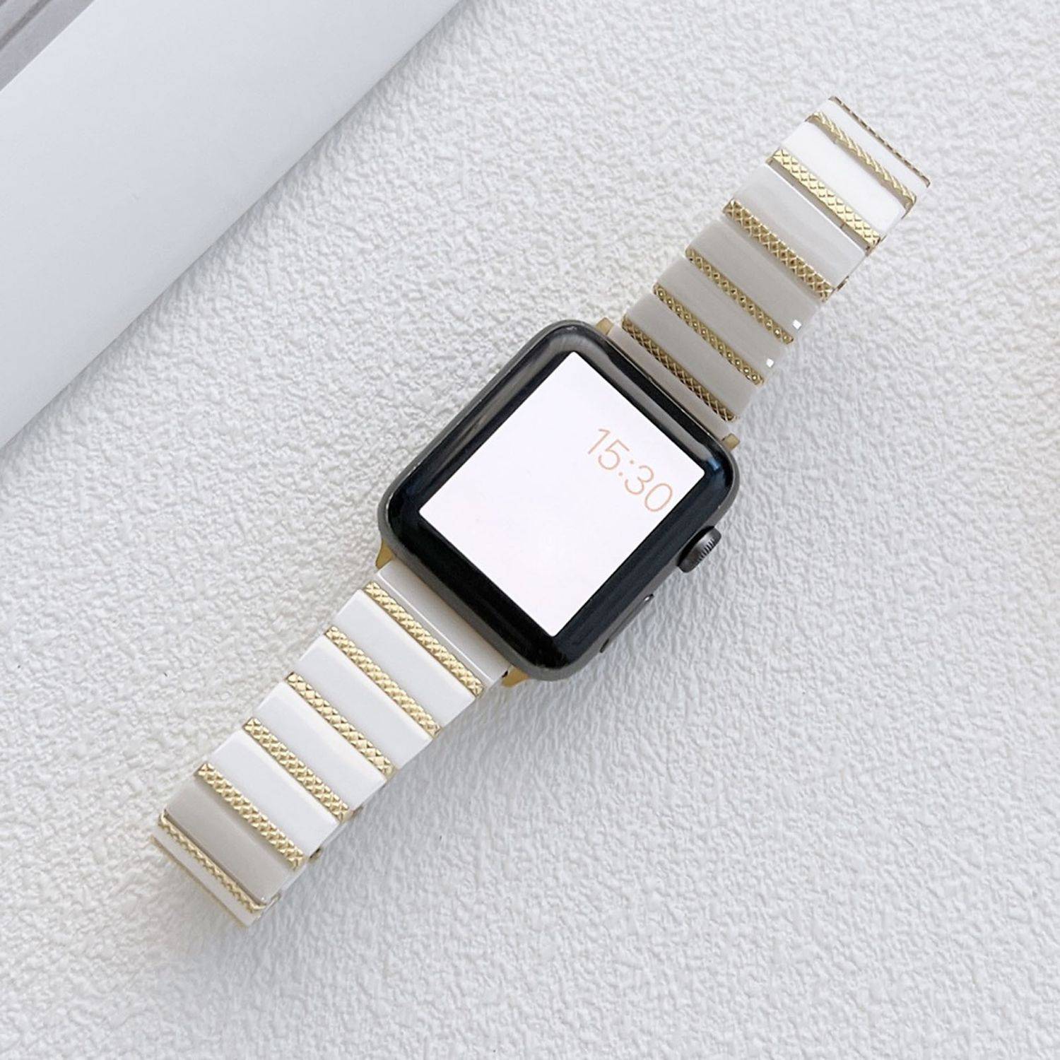 Smartwatch mit schwarzem quadratischem Display und weiß-goldgestreiftem Armband auf einer leicht strukturierten Oberfläche, Bildschirm zeigt 15:30.