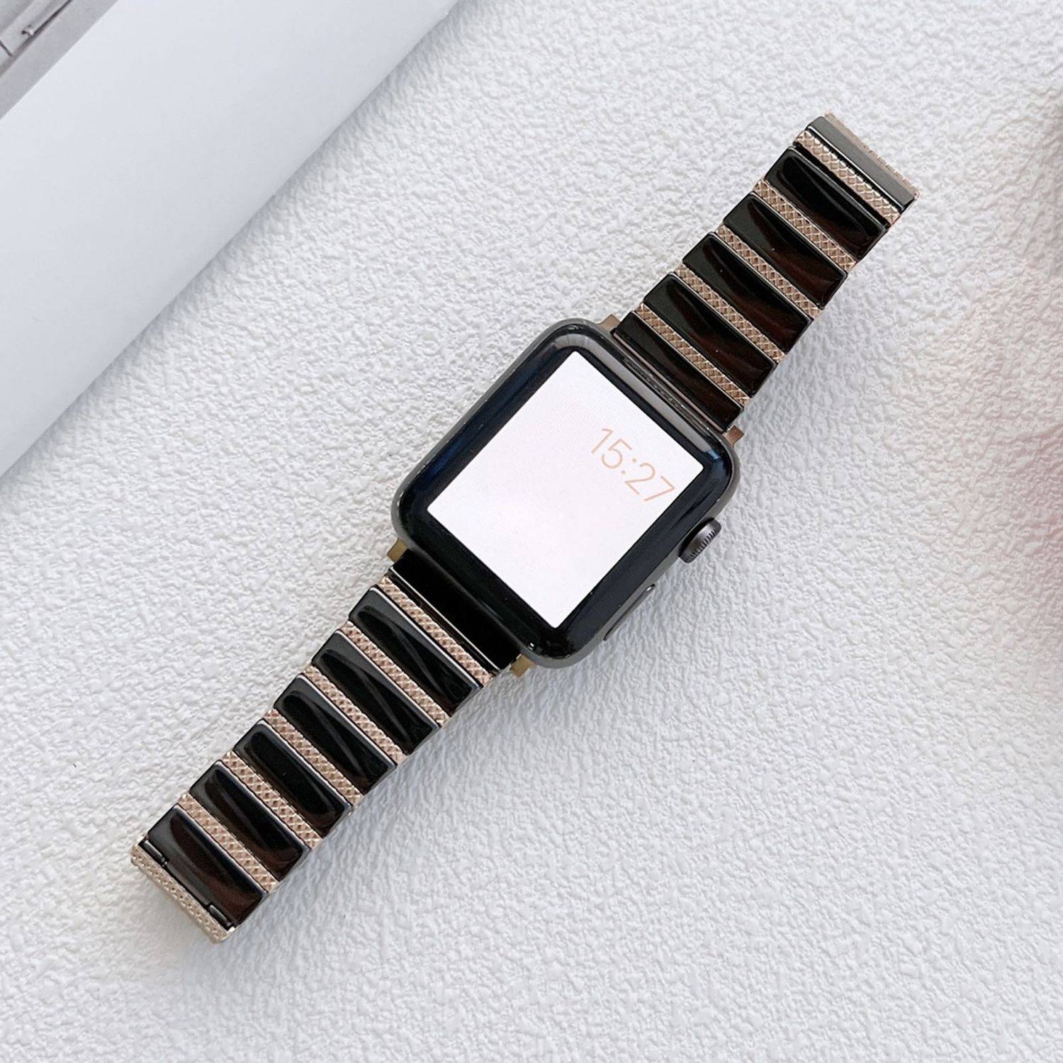 Schwarze Smartwatch mit gestreiftem Metallarmband, zeigt die Zeit 15:27 auf einem weißen Bildschirm an, liegt auf einer hellstrukturierten Oberfläche.