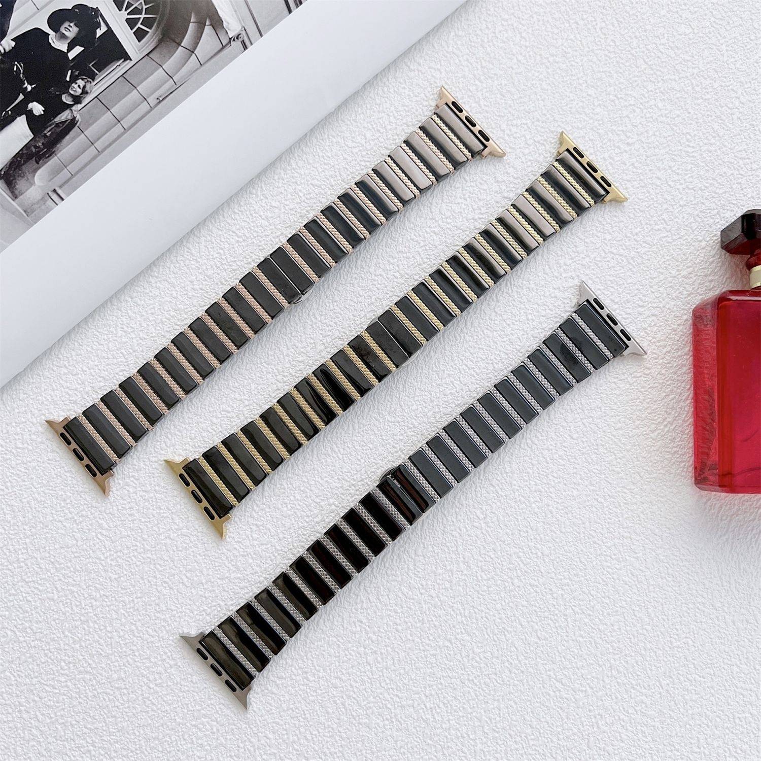 Drei metallische Uhrenarmbänder sind diagonal auf einer weißen strukturierten Oberfläche neben einer roten Parfümflasche und einem teilweise sichtbaren schwarz-weißen Foto angeordnet.