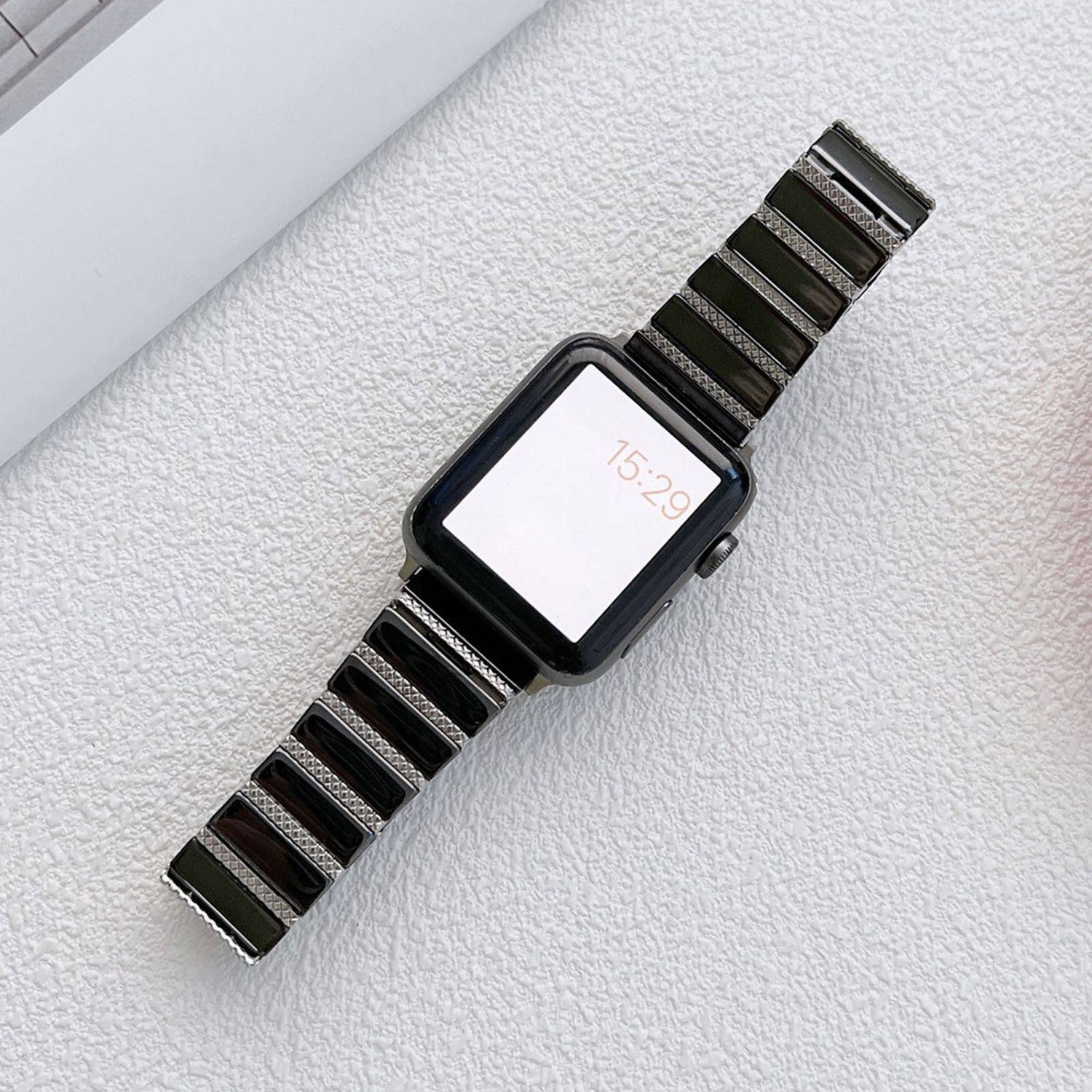 Schwarze quadratische Smartwatch mit gestreiftem Armband, die 15:29 auf einer hell strukturierten Oberfläche anzeigt.