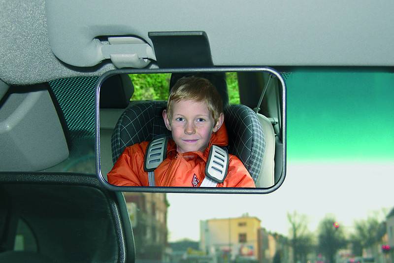 Der Rückspiegel zeigt ein Kind in einer orangefarbenen Jacke, das in einem Kindersitz auf der Rückbank eines Fahrzeugs angeschnallt ist.