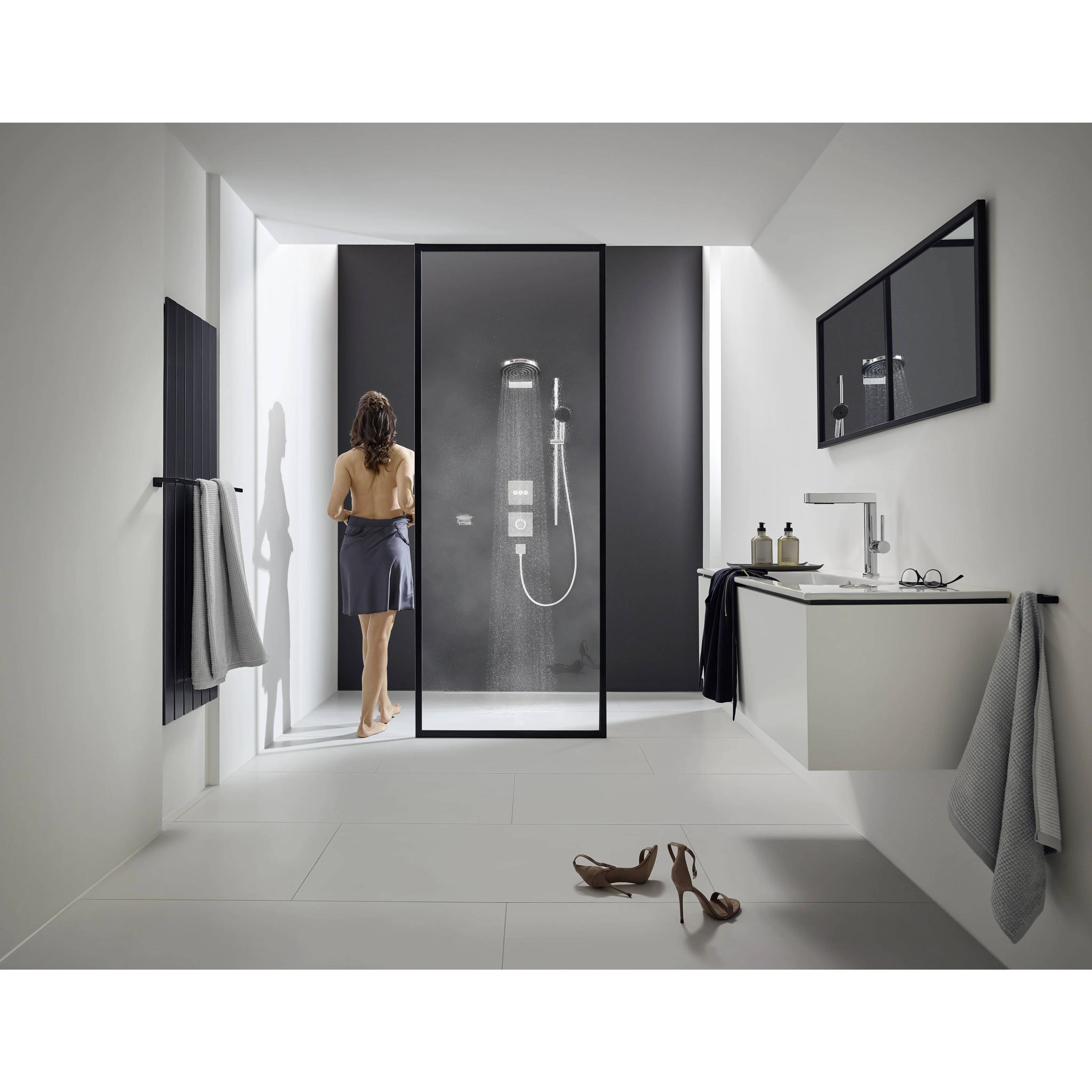 Modernes Badezimmer mit einer Frau, die neben einer gläsernen Duschkabine steht, Waschtisch und Spiegel auf der rechten Seite, hängende Handtücher und Schuhe auf dem Boden.