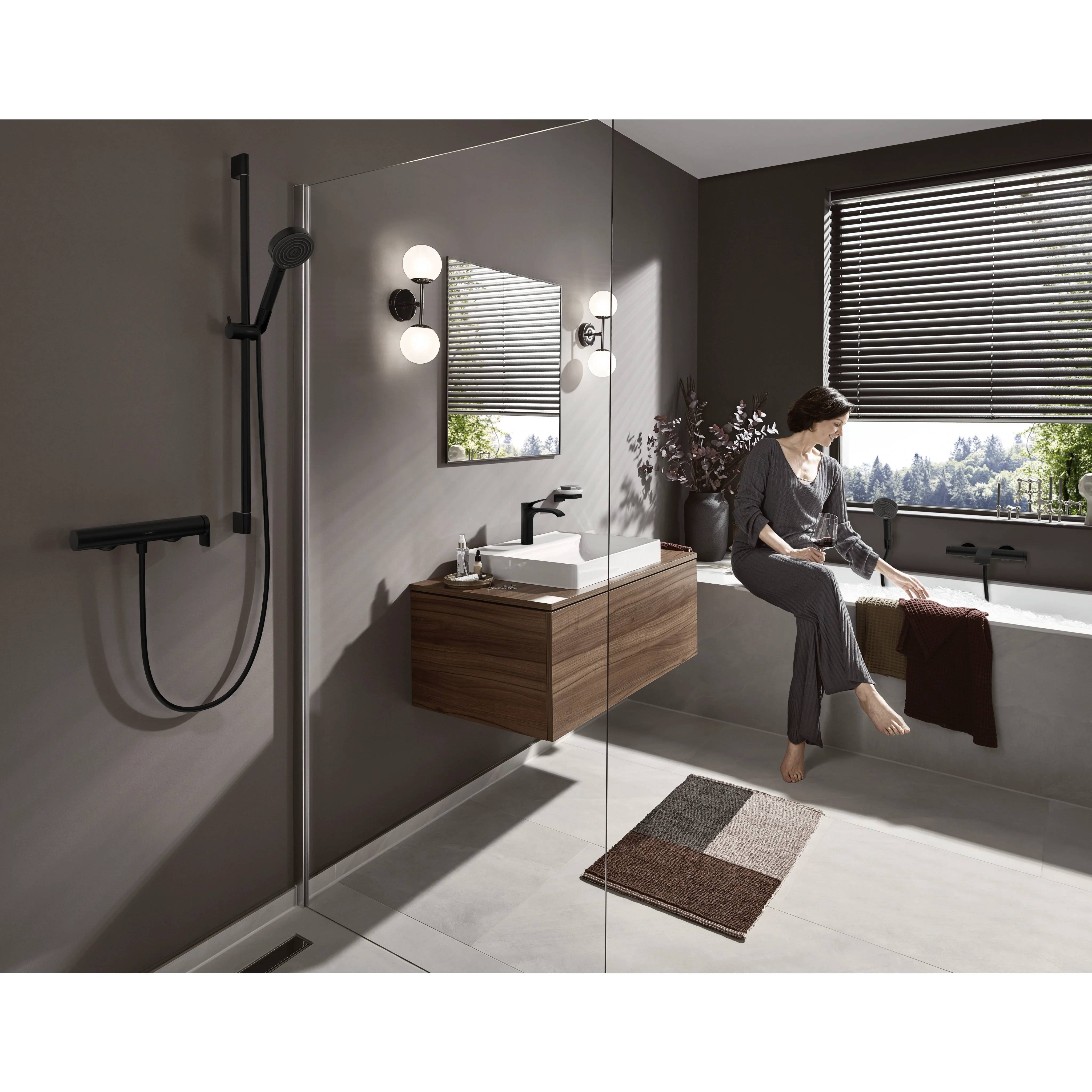 Modernes Badezimmer mit ebenem Duschbereich, Waschtisch, Badewanne an einem großen Fenster und eine Frau, die auf der Badewannenkante sitzt im weichen Tageslicht.