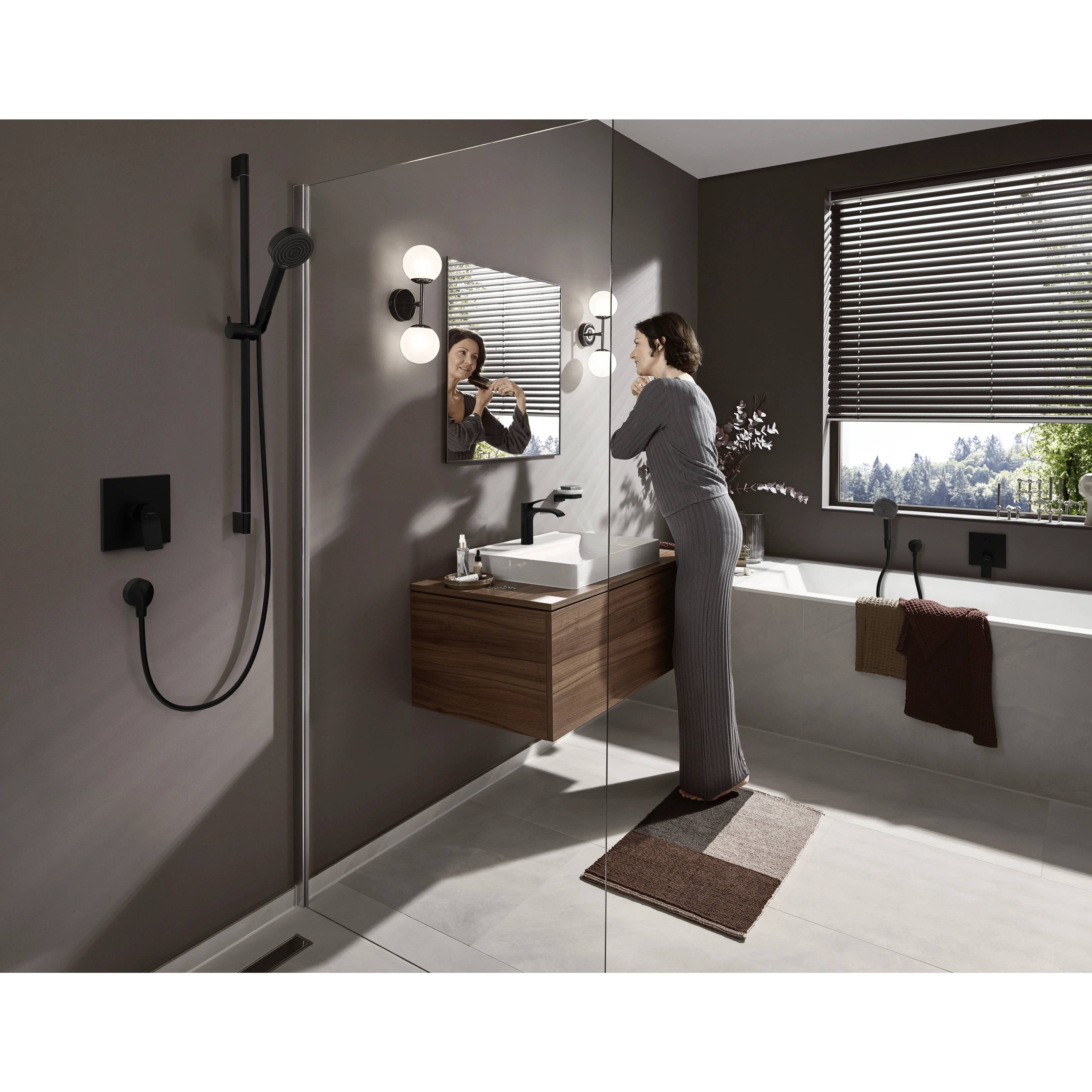 Frau, die ihre Haare in einem modernen Badezimmer neben einem Waschbecken, einer begehbaren Dusche und einer Badewanne in der Nähe eines großen Fensters bürstet.
