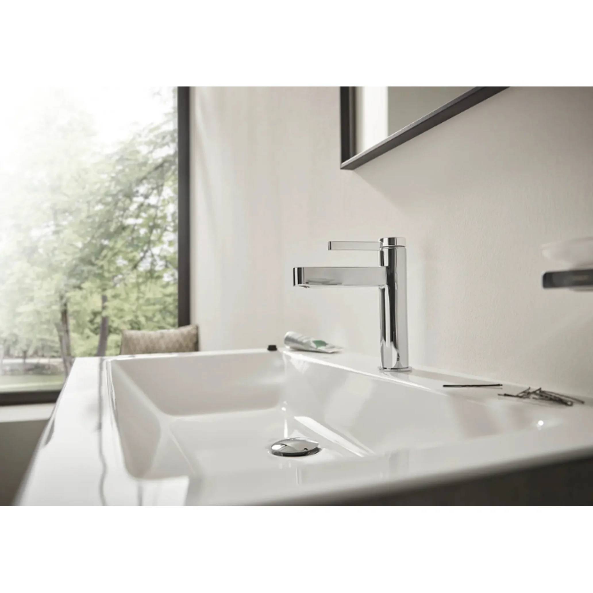 Modernes Badezimmerbecken mit verchromtem Wasserhahn neben einem Fenster mit Blick auf Bäume, in einem sauberen minimalistischen Innenraum.