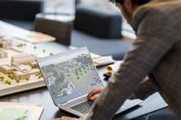 Ein Mann überprüft einen Laptop-Bildschirm, auf dem ein 3D-Lageplan angezeigt wird, neben einem Tisch-Architekturmodell in einem Büro oder Studio.