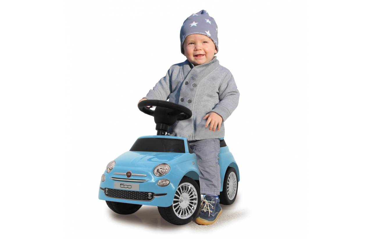 Ein lächelndes Kleinkind in einem Hut mit Sternenmuster sitzt auf einem blauen Spielzeugauto und hält das Lenkrad vor einem weißen Hintergrund.