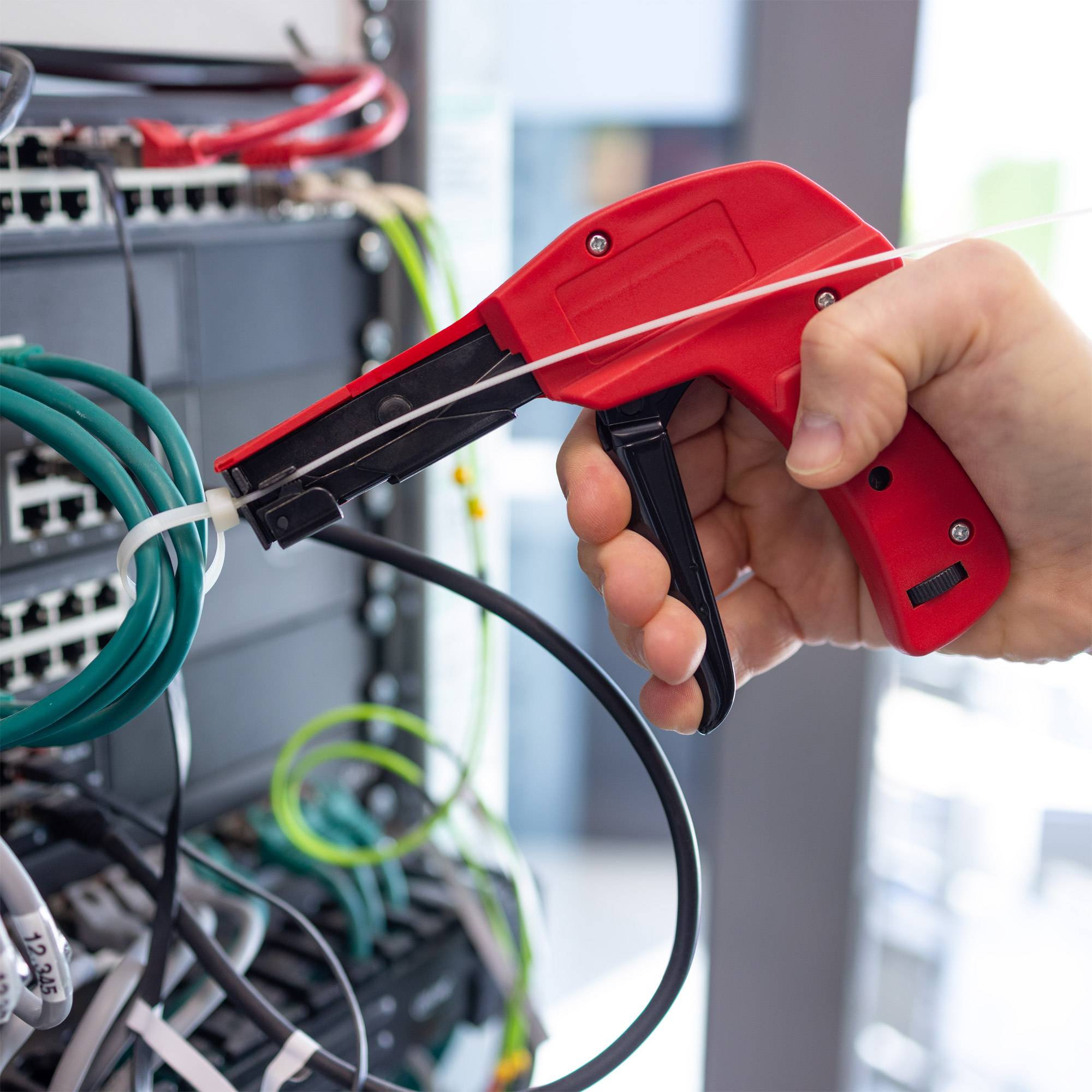 Hand mit einem roten Netzwerk-Kabeltester auf einem Ethernet-Kabel, das in einen Server oder einen Netzwerk-Switch eingesteckt ist, mit anderen gebündelten Kabeln im Hintergrund.