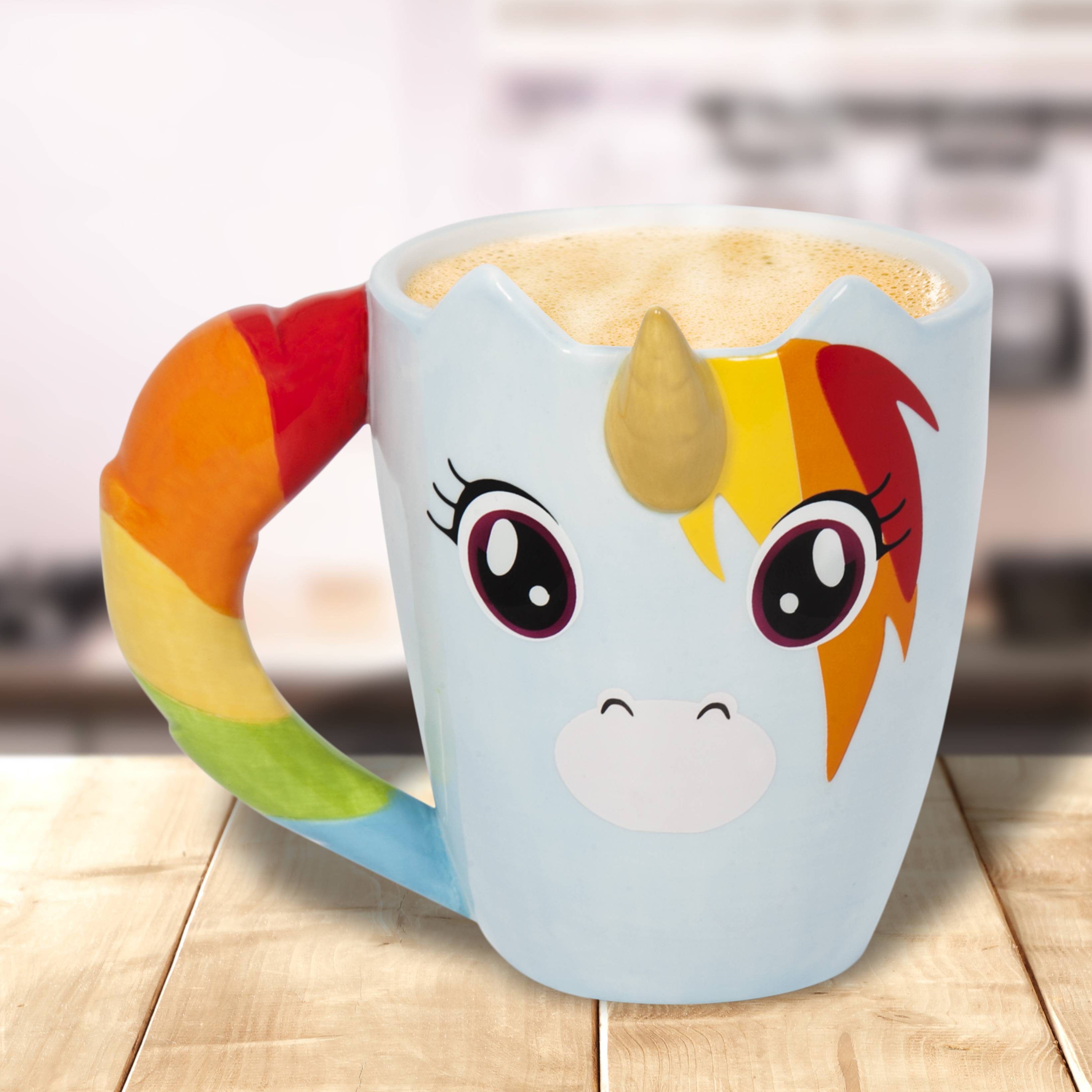 Eine hellblaue Einhorn-Tasse mit Regenbogenhenkeln und Gesichtsdesign steht auf einem Holztisch und ist mit einem schaumigen Getränk gefüllt.