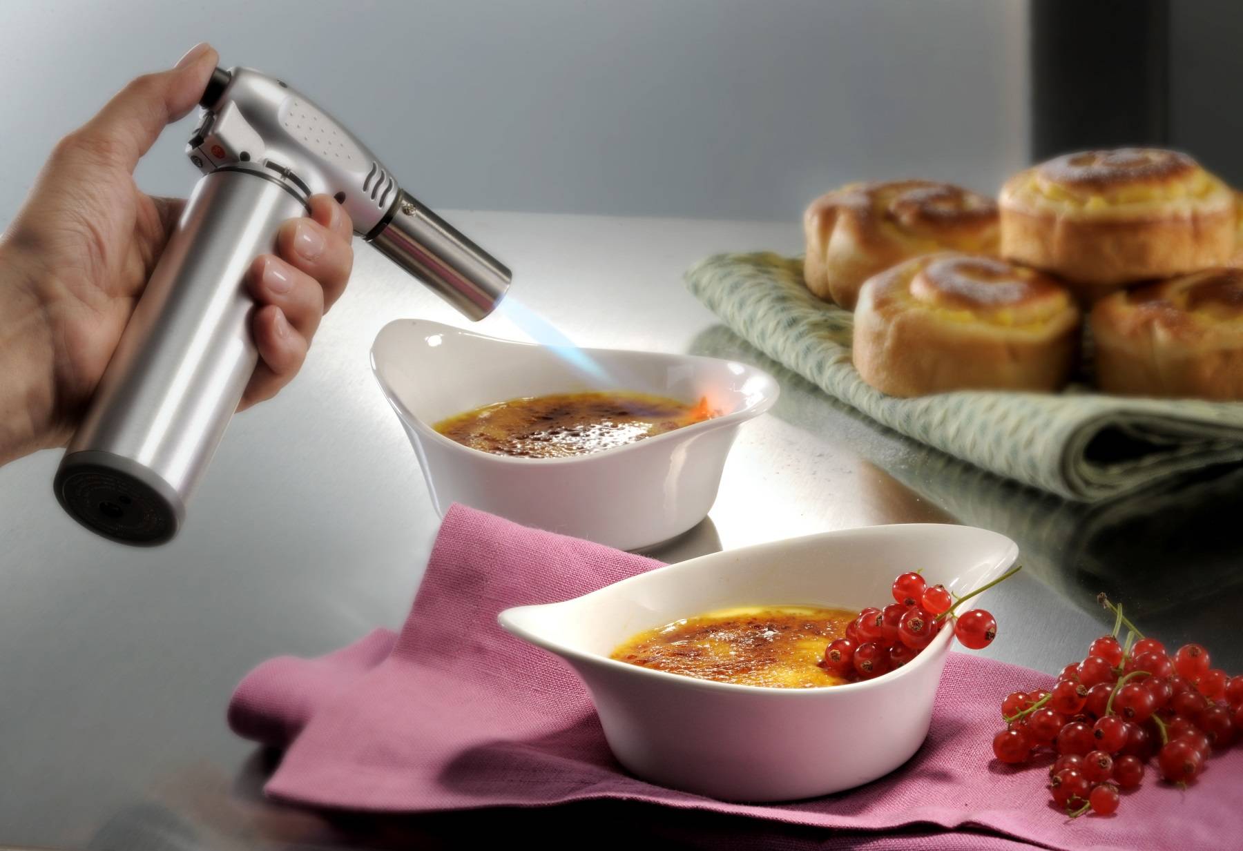 Eine Hand nutzt einen Küchenbecher, um die Oberseite eines Desserts in einer weißen Schale zu karamellisieren, mit Gebäck und roten Johannisbeeren daneben.