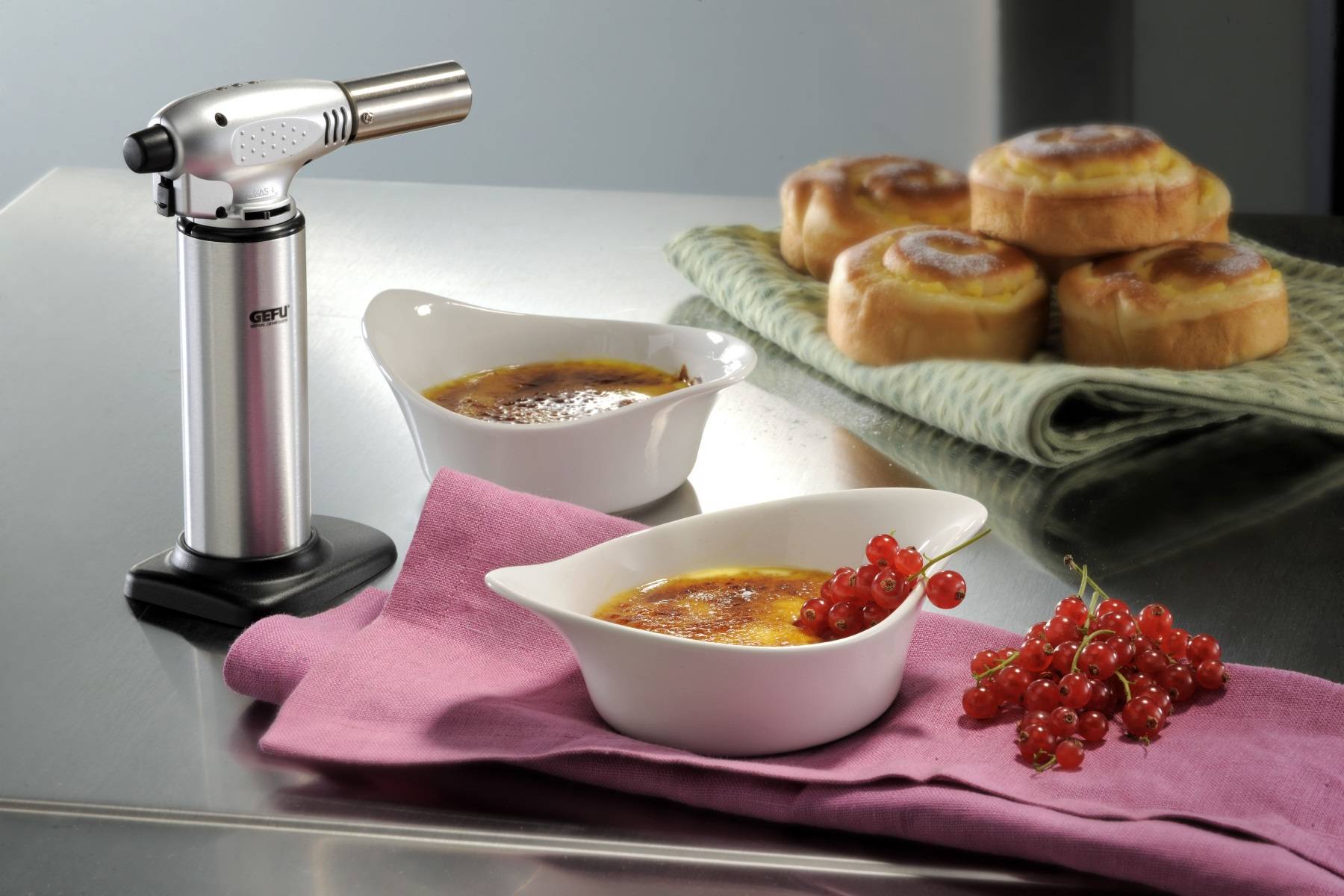 Küchenfeuerzeug neben zwei Crème-Brûlée-Schalen mit karamellisierten Oberseiten, roten Johannisbeeren und gebackenen Desserts im Hintergrund auf einer Arbeitsplatte.