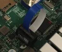 Nahaufnahme einer Leiterplatte mit einem blau-weißen Flachbandkabel, das mit einem schwarzen Bauteil verbunden ist.