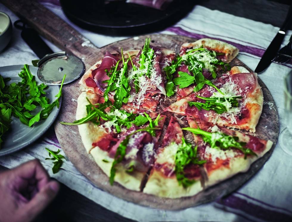 Rustikale Pizza, belegt mit hauchdünn geschnittenem Fleisch, Grünzeug und geriebenenem Käse auf einem Holzbrett, mit zusätzlichen Kräutern und einem Pizzamesser daneben.