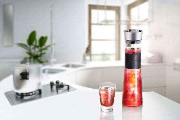 Hohes Glaskaraff und kleines Glas mit eisgekühltem Rotfruchtsaft auf einer Küchenarbeitsplatte, mit einem Mixer im Hintergrund.