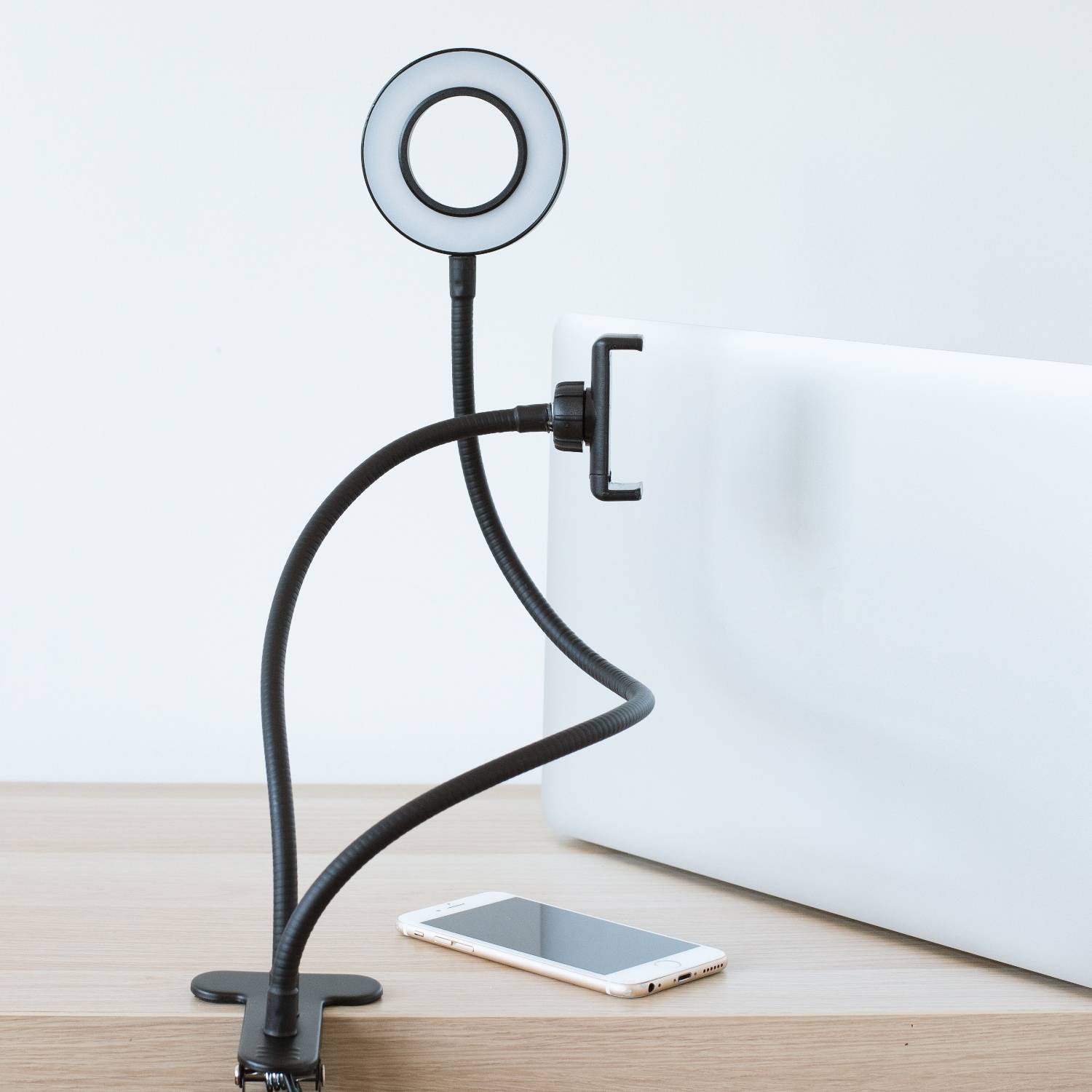 Ringlicht-Telefonhalter-Klemme an einem Schreibtisch befestigt, mit einem Smartphone, das auf dem Tisch neben einem weißen Monitor oder Panel liegt.