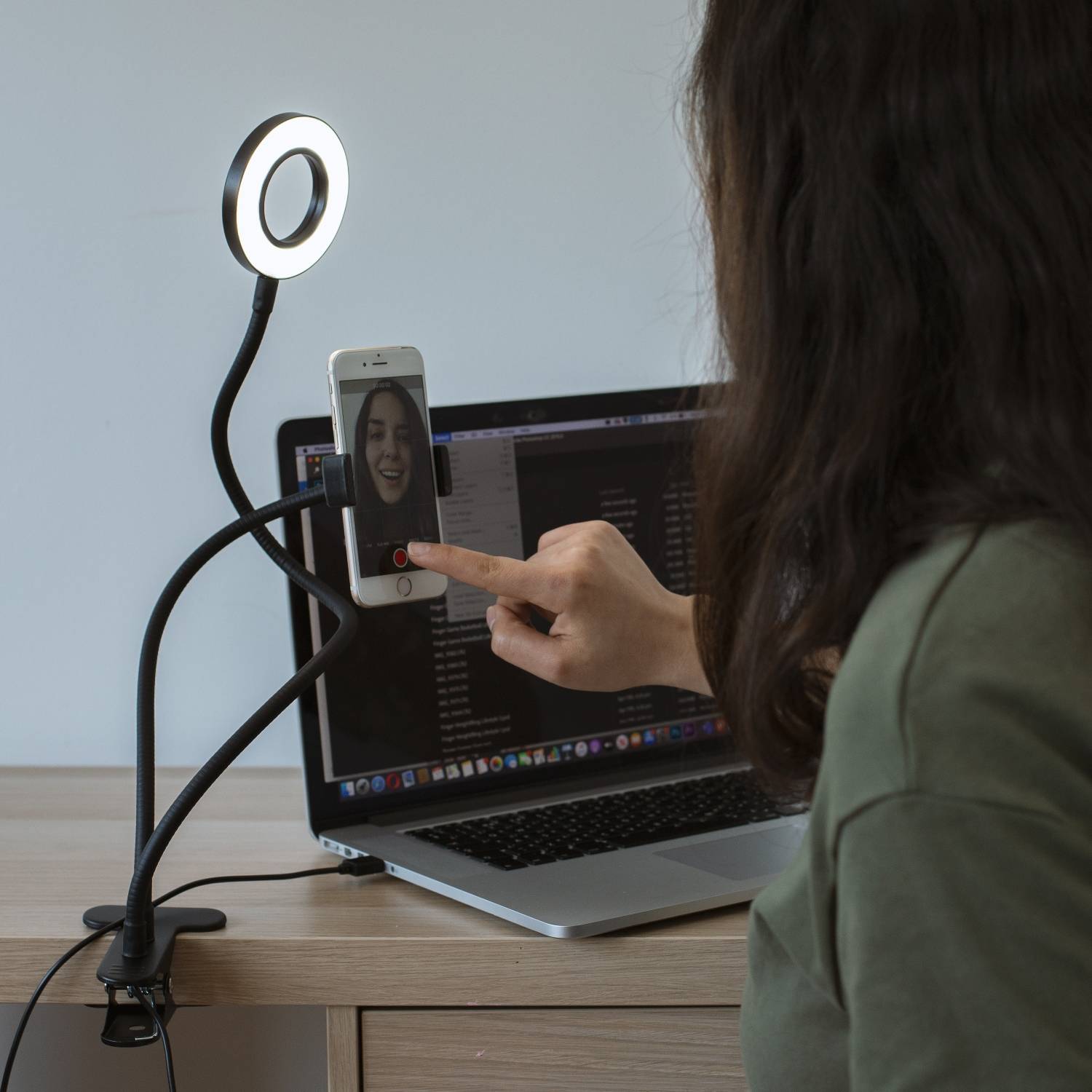 Eine Frau nutzt ein Smartphone auf einem Ringlicht-Stativ, um einen Videoanruf auf einem Laptop zu führen, während ihr Finger den Smartphone-Bildschirm berührt.
