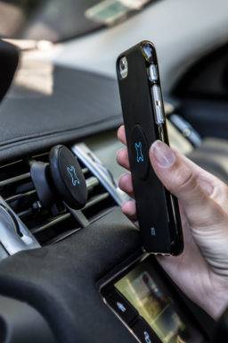 Hand hält ein Smartphone in der Nähe einer Telefonhalterung im Autolüftungsschlitz des Armaturenbretts, was auf Navigation im Auto oder freihändige Nutzung hinweist.