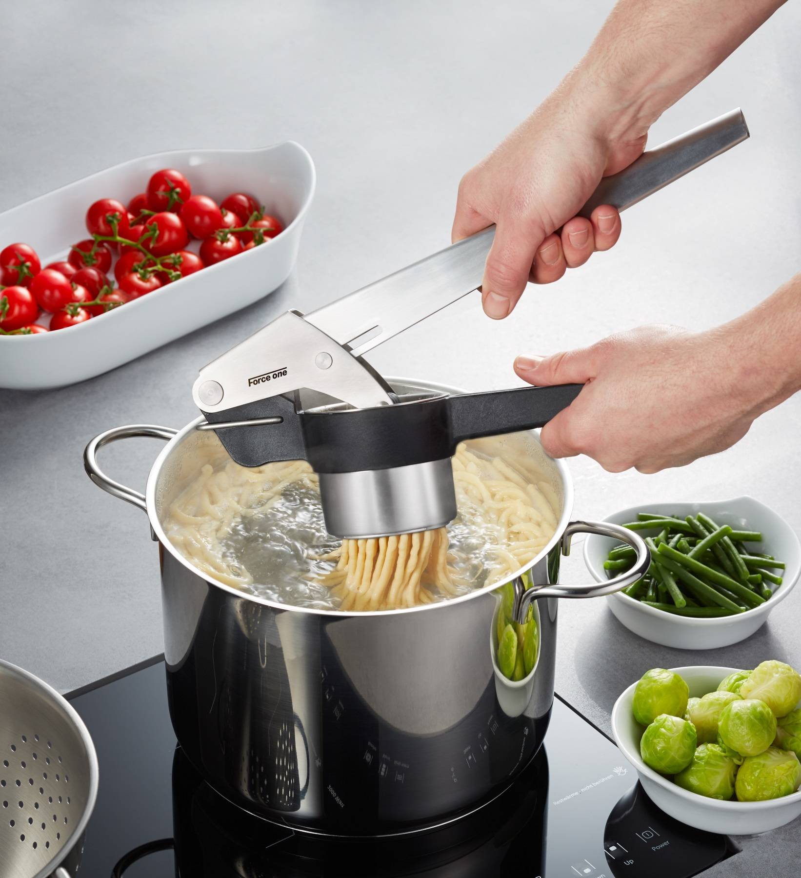 Hände, die eine Kartoffelpresse verwenden, um gekochte Nudeln in einen Topf zu drücken, mit Schüsseln mit Kirschtomaten, grünen Bohnen und Rosenkohl in der Nähe.