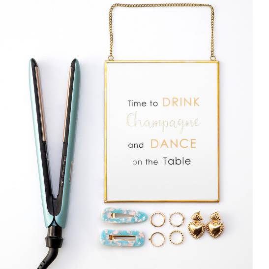 Goldgerahmtes Wandschild mit der Aufschrift „Time to DRINK Champagne and DANCE on the Table" neben einem türkisfarbenen Haarglätter und kleinen Ohrringen.