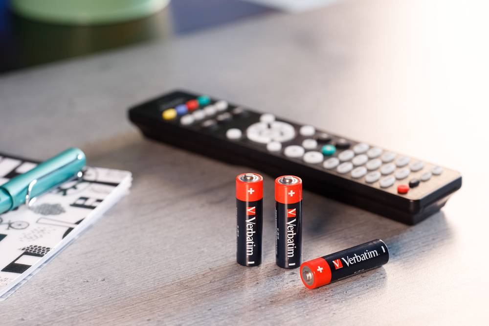 Drei Verbatim AA-Batterien auf einem Tisch vor einer Fernbedienung eines Fernsehers.