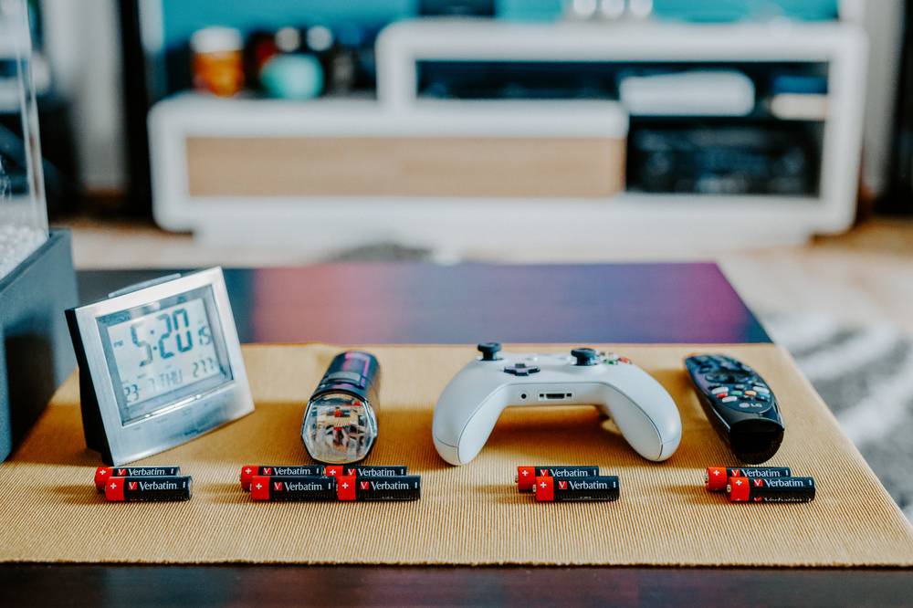 Eine Tischplatzierung mit einer digitalen Uhr, einem Spielzeugfahrzeug, einem Spielecontroller, einer Fernbedienung für den Fernseher und Verbatim-Batterien, die in einer Reihe auf einem Holztablett angeordnet sind.