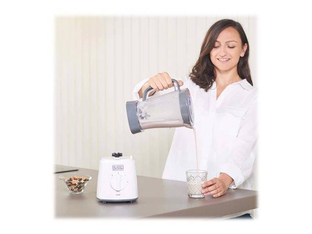 Eine Frau gießt einen Smoothie aus einem Blenderkrug in ein Glas auf einer Küchenarbeitsplatte. Eine Schüssel mit Nüssen steht daneben.