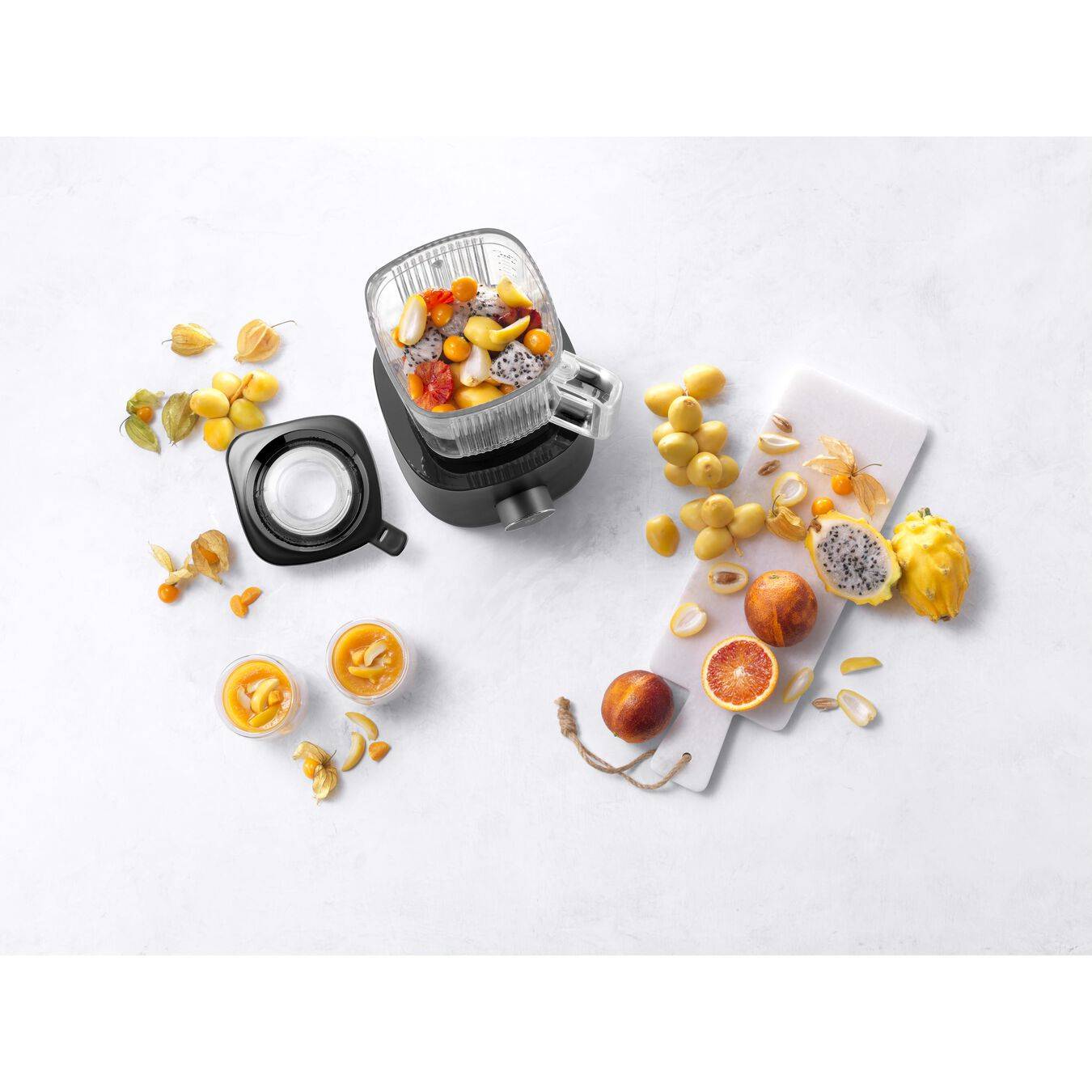Draufsicht auf einen Mixer mit gemischten Fruchtzutaten im Behälter, mit geschnittenem Obst und einem abgenommenen Deckel auf einer weißen Oberfläche.
