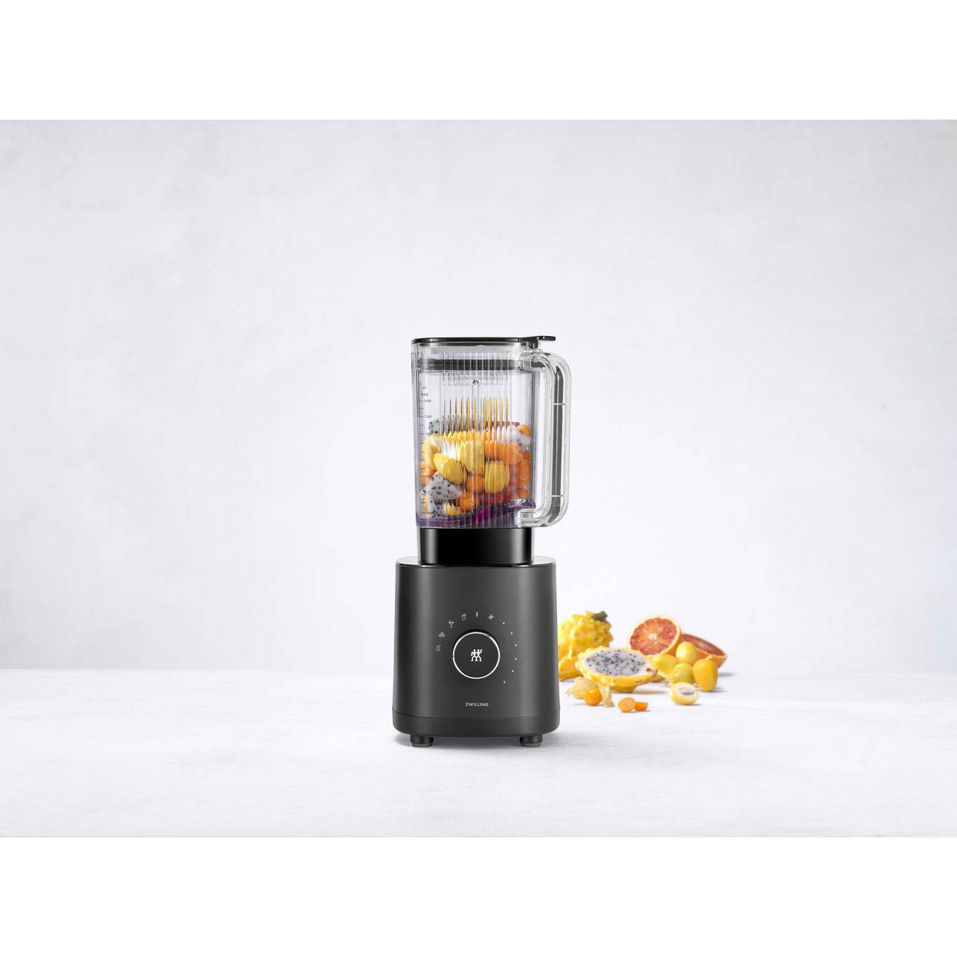 Ein schwarzer Standmixer mit einem durchsichtigen Behälter gefüllt mit Fruchtstücken, mit zerkleinertem Obst und Zutaten, die daneben auf weißem Hintergrund verstreut sind.