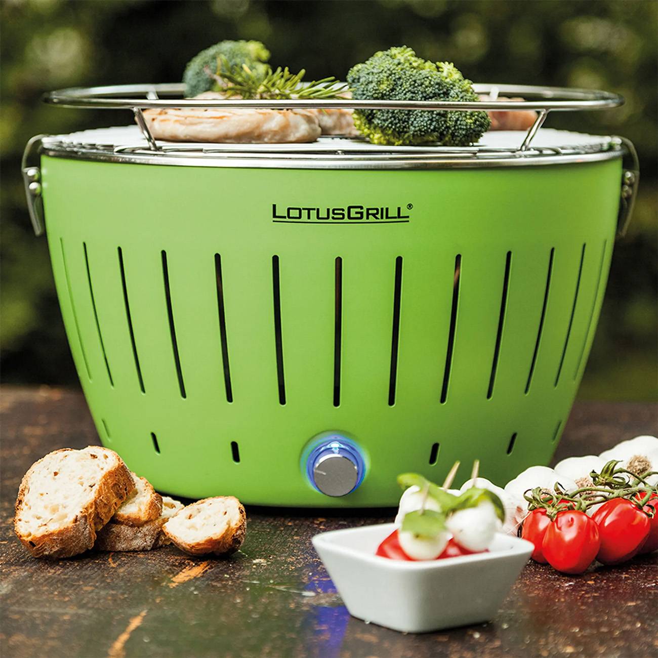 Grüner LotusGrill Tischgrill mit Holzkohle und Lebensmitteln zum Kochen oben, umgeben von Brot, Gemüse und Tomaten im Freien.