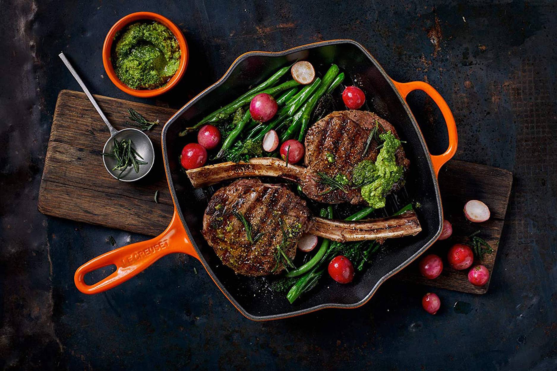 Vogelperspektive einer Gusseisen-Bratpfanne mit gegrillten Steaks, grünen Bohnen, Radieschen und Kräutern auf einem Holzbrett, mit Sauce und Besteck in der Nähe.