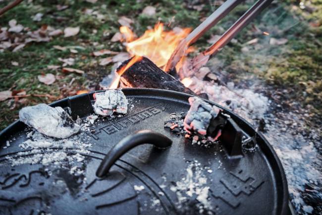 Zangen heben glühende Kohlen aus einem Lagerfeuer neben einem gusseisernen Dutch Oven mit Deckel auf einem Campingplatz im Freien.