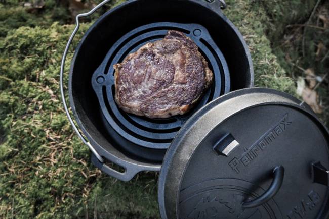 Ein angebratenes Steak gart in einem gusseisernen Bräter über Gras, wobei der Deckel daneben ruht.