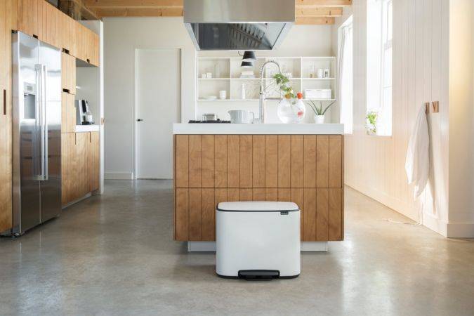 Moderne Küche mit einem weißen Tritt-Mülleimer in der Mitte vor einer Holzinsel, einem Edelstahl-Kühlschrank auf der linken Seite und hellen Fenstern auf der rechten Seite.