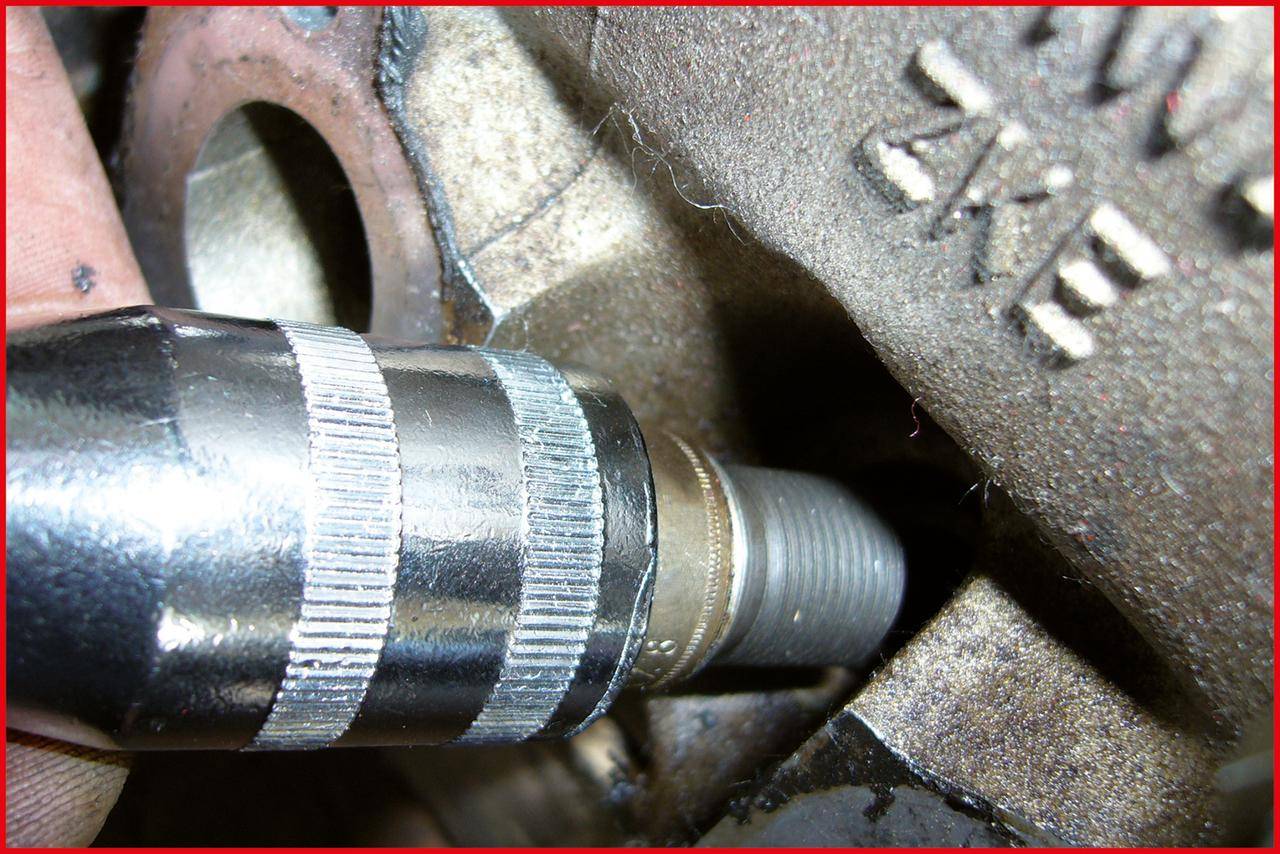 Eine Nahaufnahme einer Zündkerze, die teilweise in einen Motorzylinderblock eingeschraubt ist und dabei das Metallgehäuse und das Gewinde der Kerze hervorhebt.