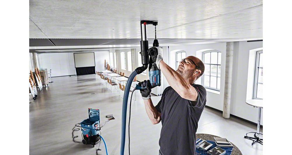 Eine Person mit Brille bedient ein Deckenbohrgerät in einem großen, leeren Raum mit Reihen von Stühlen und einem Tisch, was auf Einrichtungs- oder Wartungsarbeiten hindeutet.
