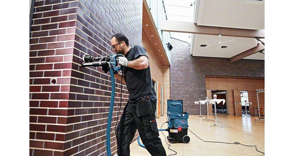Eine Person in Arbeitskleidung bohrt mit einem Elektrowerkzeug, das an ein Absaugsystem angeschlossen ist, in eine Ziegelwand im Innenbereich.