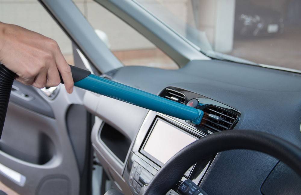 Eine Person verwendet ein blaues Reinigungswerkzeug, um eine Lüftungsöffnung im Wageninneren abzustauben.