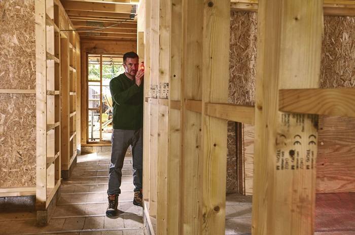 Eine Person in einem teilweise fertiggestellten Holzgebäude verwendet eine Wasserwaage, um die Ausrichtung einer Wand zu überprüfen. Sonnenlicht strömt durch ein Fenster.