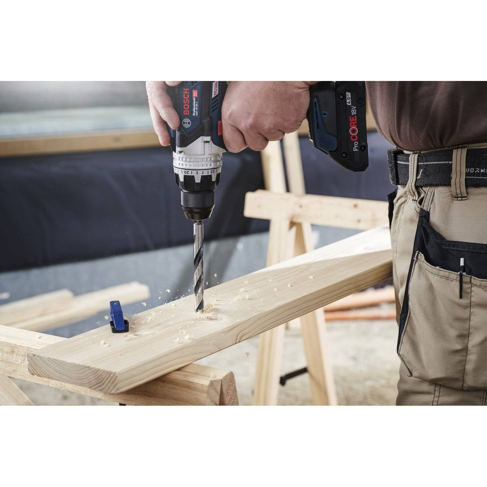 Eine Person, die einen Bosch-Bohrer verwendet, um ein Loch in ein Holzbrett auf einer Werkbank zu bohren. Der Bohrer ist mitten in der Operation, mit sichtbaren Holzspänen.