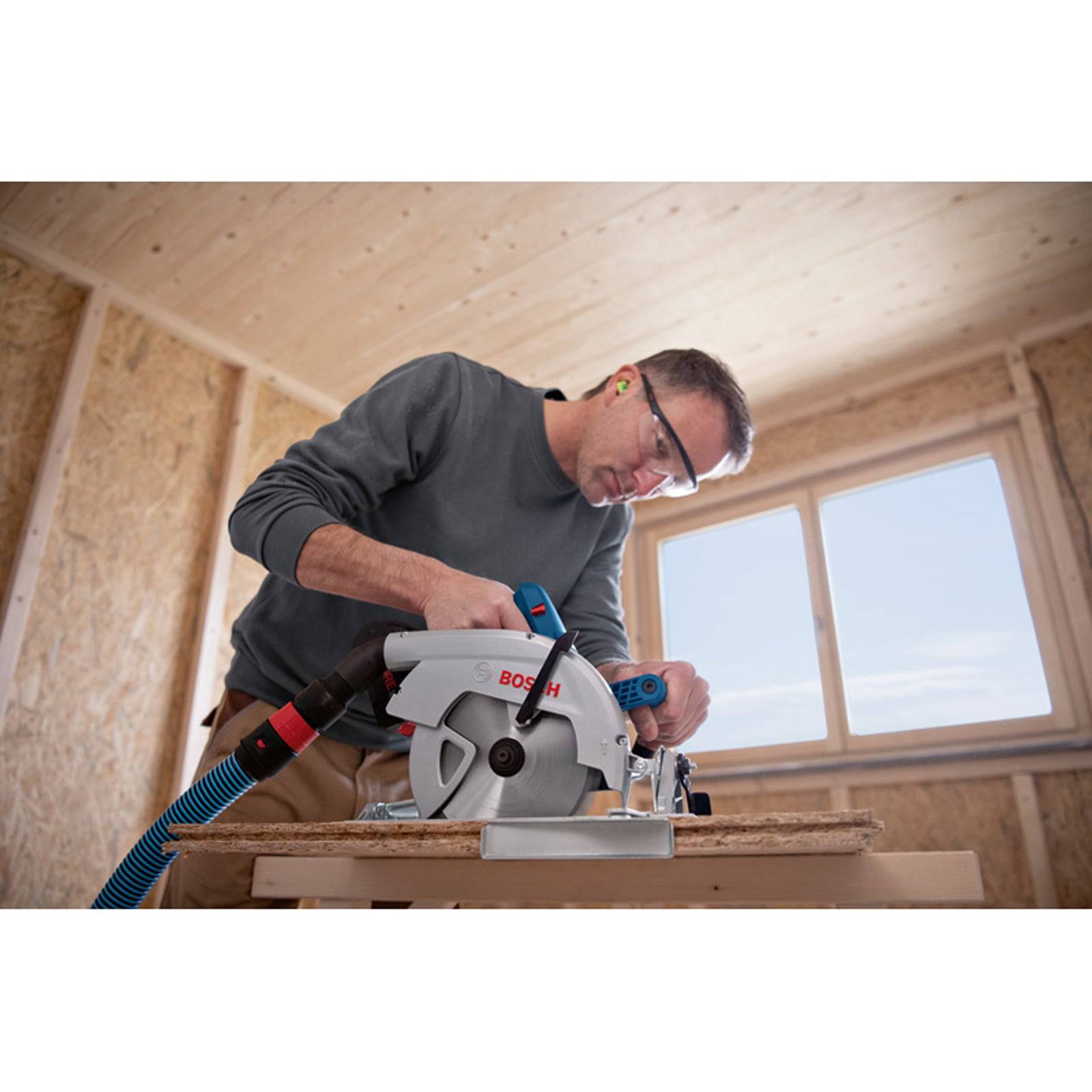 Ein Mann in Schutzausrüstung benutzt eine Bosch-Kreissäge auf Holz im Inneren eines teilweise fertiggestellten Holzgebäudes, wobei Sonnenlicht durch ein Fenster fällt.
