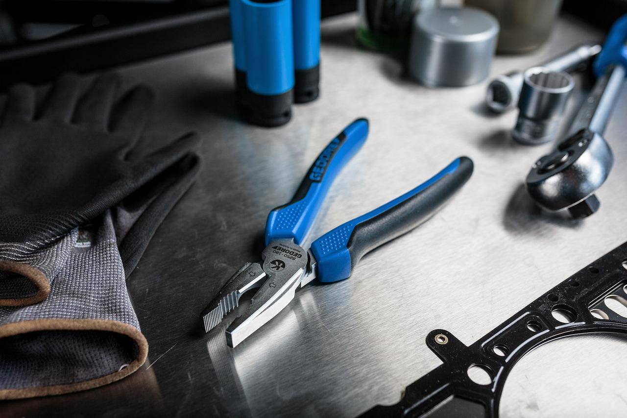 Blaugriffige Zange auf einer metallischen Oberfläche mit Handschuhen in der Nähe, daneben Rohre und zylindrische Objekte verschwommen im Hintergrund.