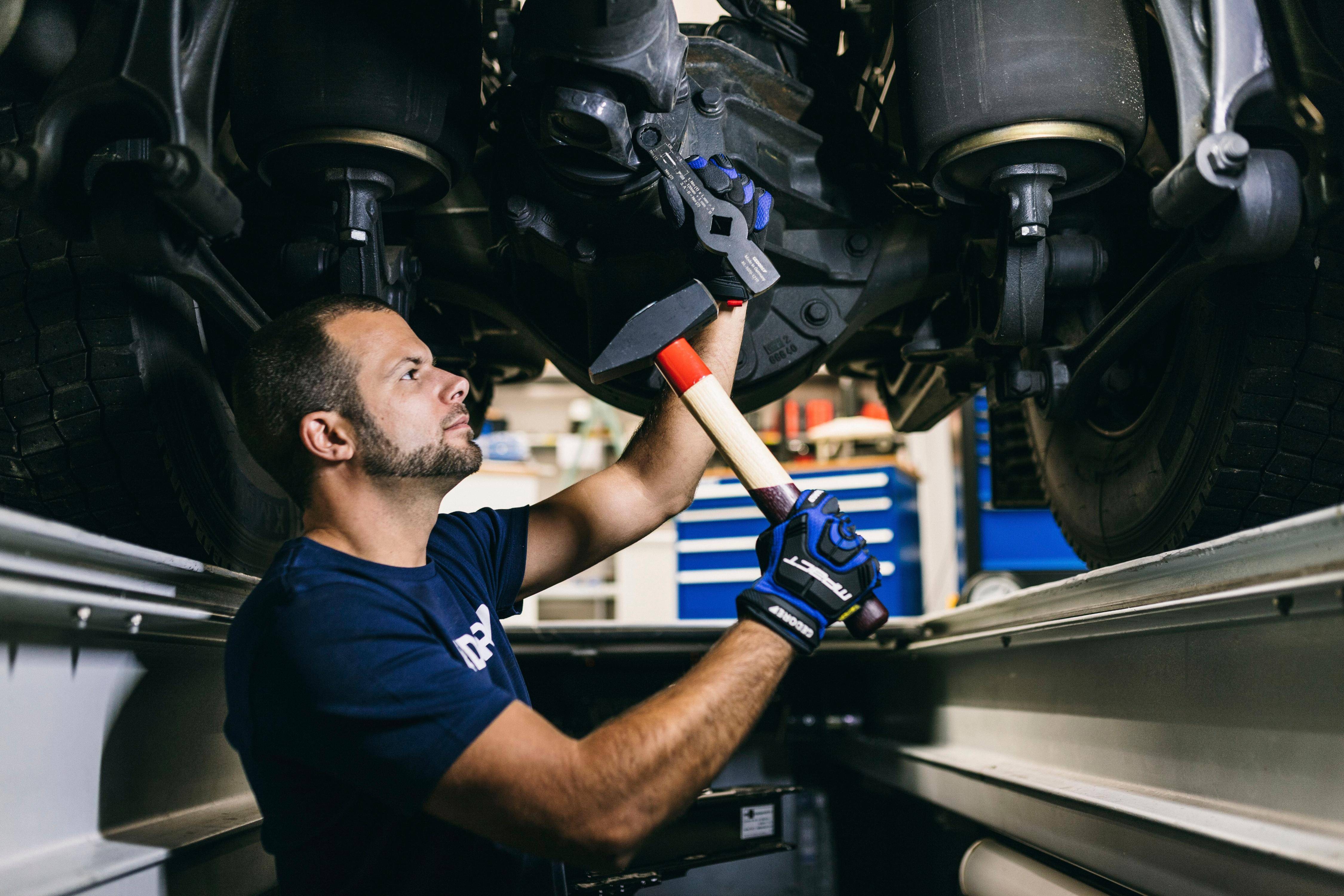 Ein Mechaniker in einem blauen Hemd und mit Handschuhen repariert die Unterseite eines Busses in einer Werkstatt und nutzt ein Werkzeug, um ein Teil zu justieren.